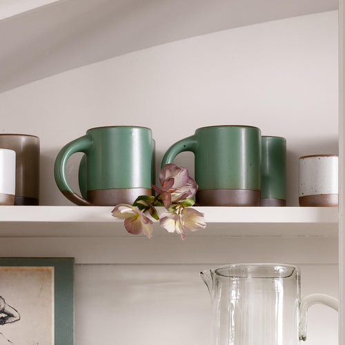 Set of green mugs on a shelf with flowers and a glass pitcher.