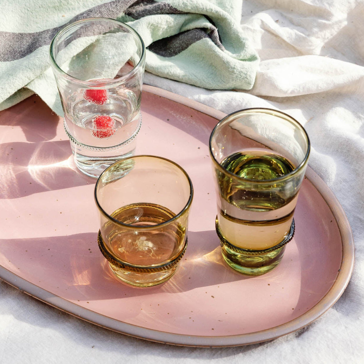 Pink oval serving platter holding three banded clear and green drinking glasses on a picnic blanket beside a striped tea towel.