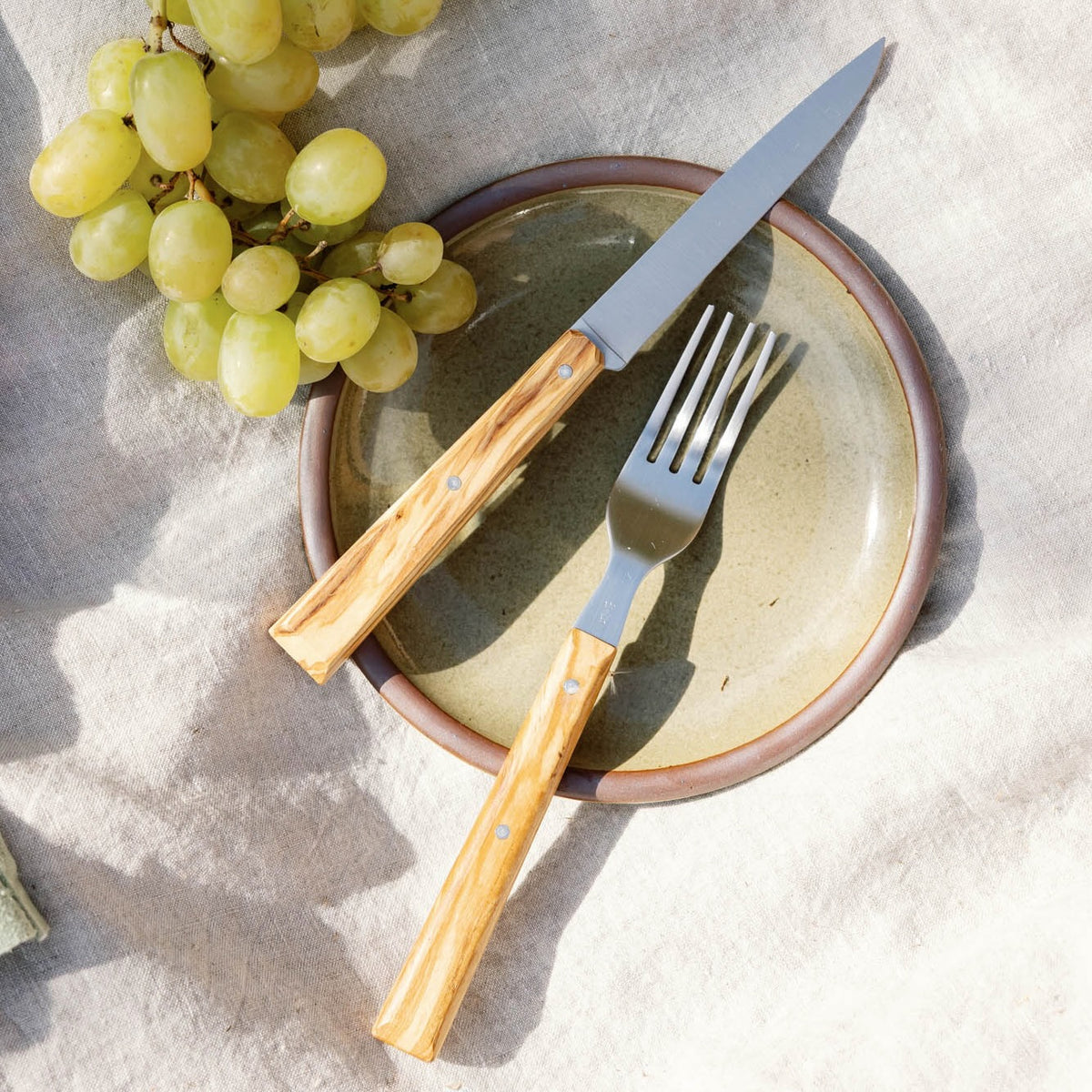 Knife and fork with wooden handles resting on a ceramic plate beside green grapes on a linen surface.