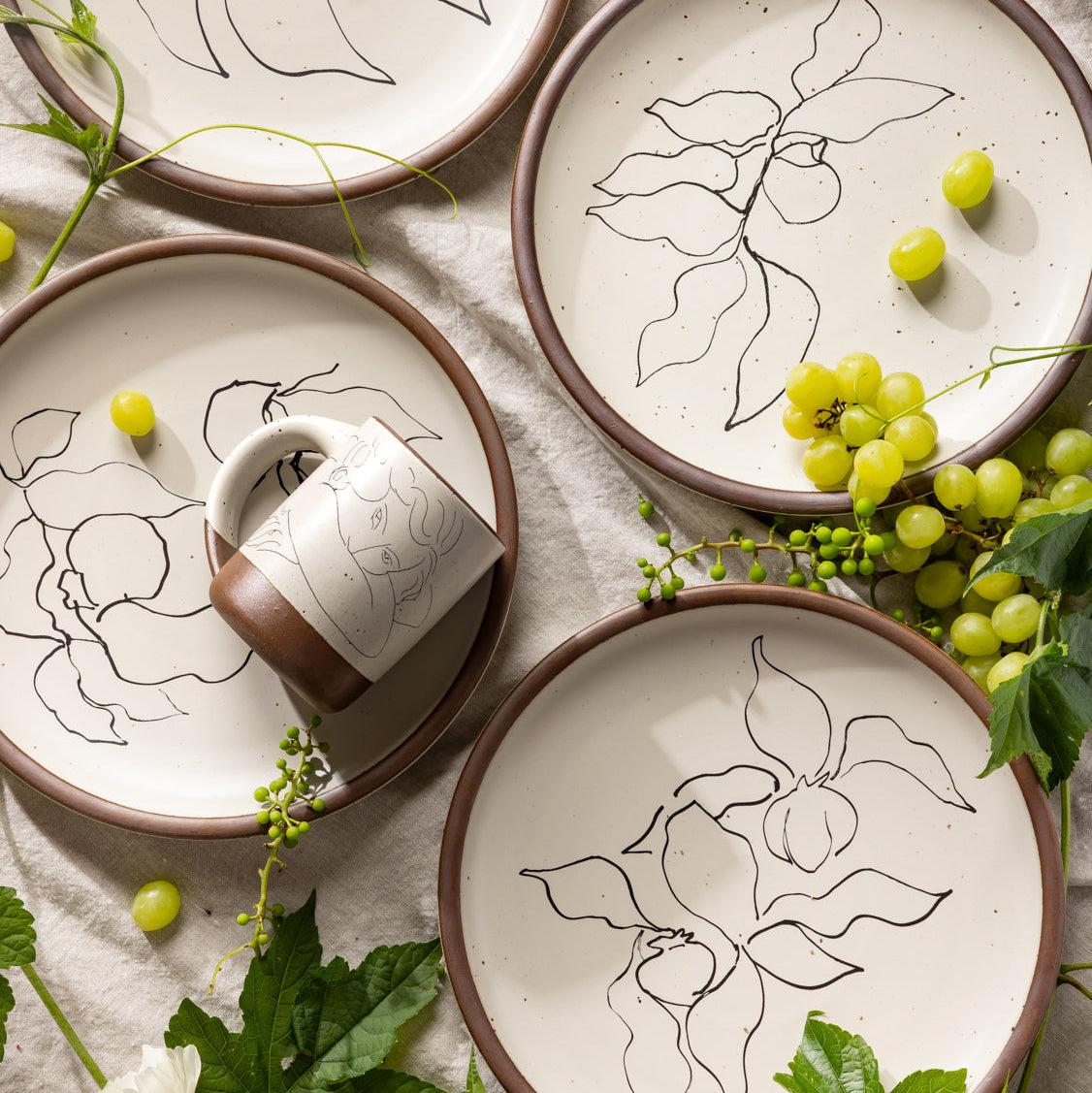 An overhead view of Dinner Plates and The Mug featuring line drawings by Henri Matisse styled with greenery and grapes.