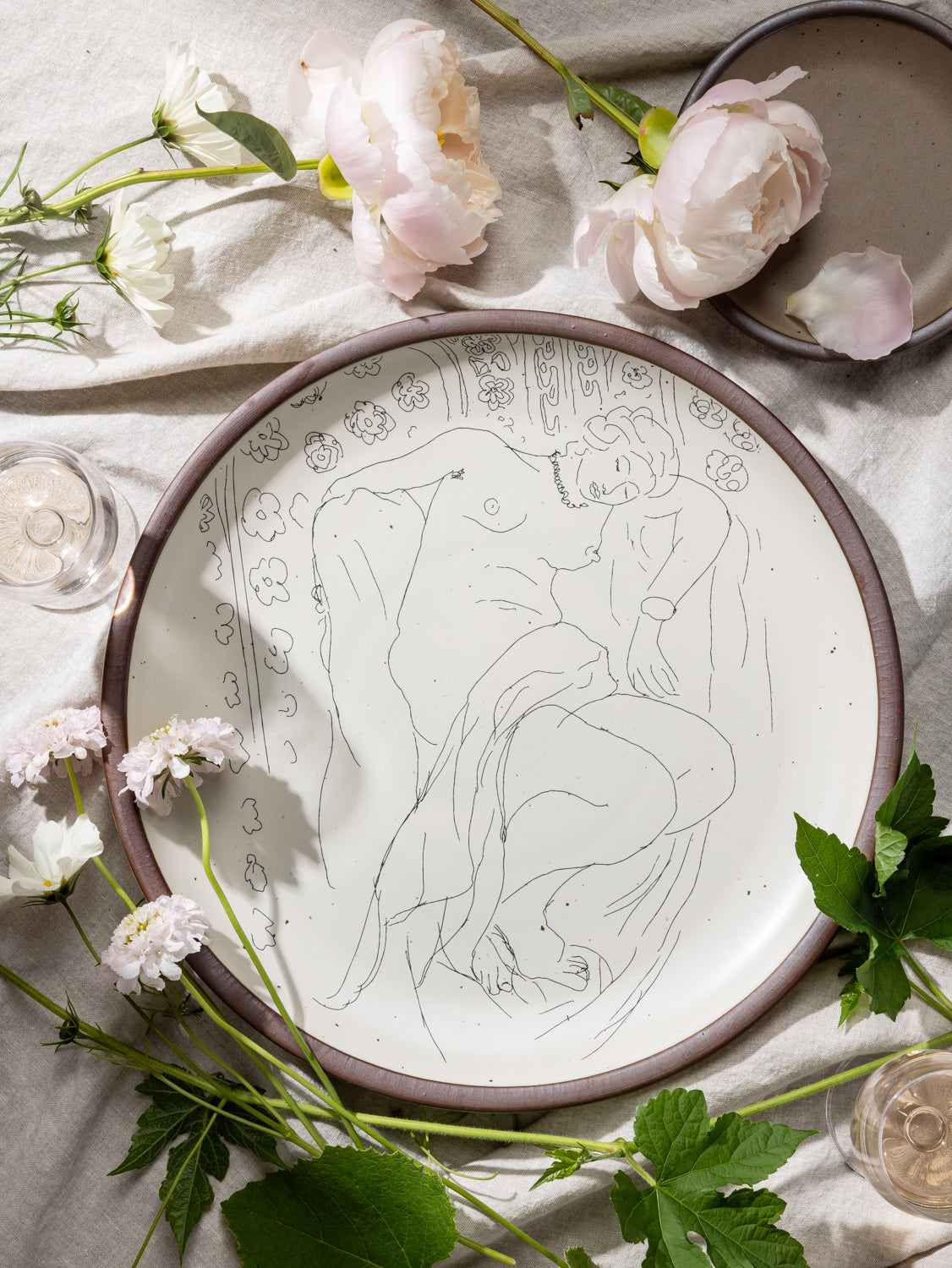A large serving platter in a warm off-white color with unglazed rim with a black decal of the artwork, Figure endormie sur fond moucharabieh, from Henri Matisse, styled on a tablecloth with grapes and flowers.