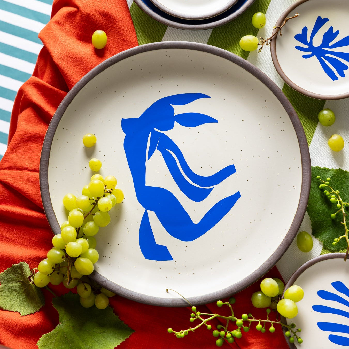 A styled flat-lay of ceramic tableware on a bright orange-red tablecloth, featuring white plates and bowls with deep blue interiors and decals of Henri Matisse's accented withgrapes