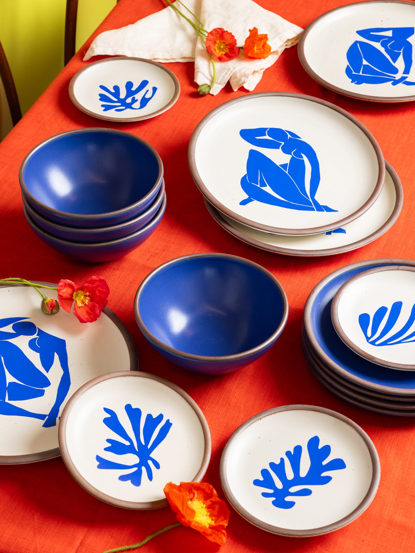 A styled flat-lay of ceramic tableware on a bright orange-red tablecloth, featuring white plates and bowls with deep blue interiors and decals of Henri Matisse's accented with small red flowers.