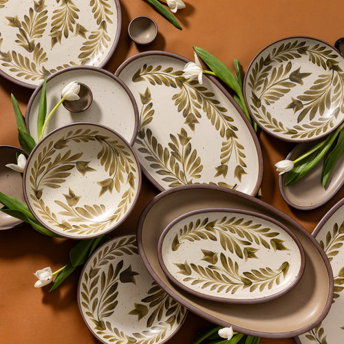 Set of ceramic platters and plates in various sizes with painted leaf patterns on artfully arranged with flowers on a brown background.