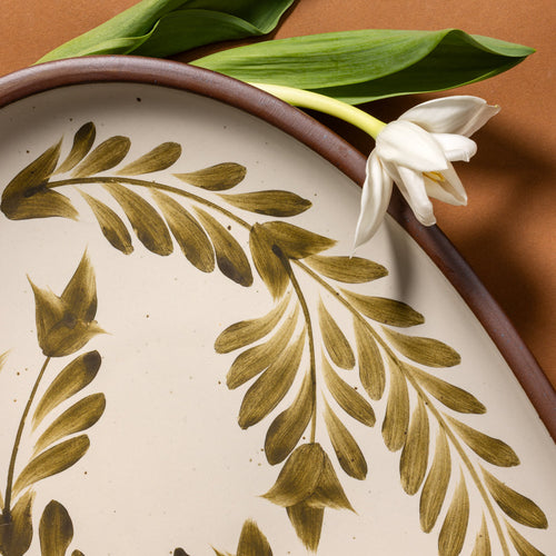 Closeup of a large oval ceramic platter in a warm, tan-toned, off-white color featuring iron speckles and an unglazed rim, with handpainted greenery in an olive green color.