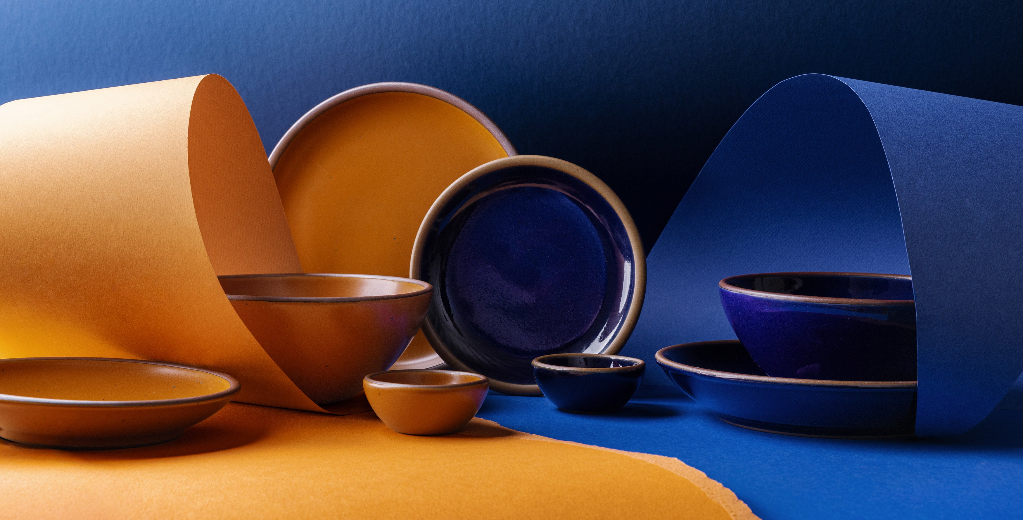 A styled studio arrangement of ceramic tableware in two contrasting glazes: a dark glossy blue and a warm earthy orange. The set includes plates, bowls, and small cups, some nested together and others displayed upright, set against a split blue and orange paper background.