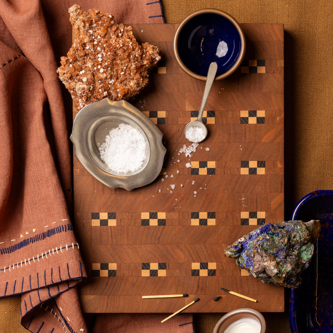 Wooden cutting board with salt, ceramic dishes, a candle, minerals, and matches arranged on rust-colored textiles.