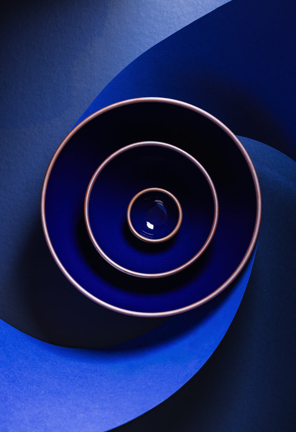 Three nested ceramic bowls in a dark glossy blue glaze with contrasting brown rims, arranged in a spiral pattern on a dark blue background.