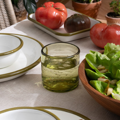 An olive green short transparent drinking glass, filled with water, on a tablescape with white enamel plates and bowls, and tomatoes and salad.