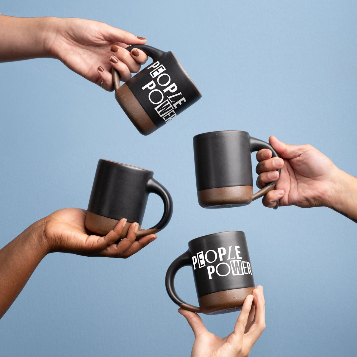 Four hands hold up ceramic mugs in a graphite black color with an unglazed base and rim, with a decal in white that reads 'PEOPLE POWER', against a blue background.