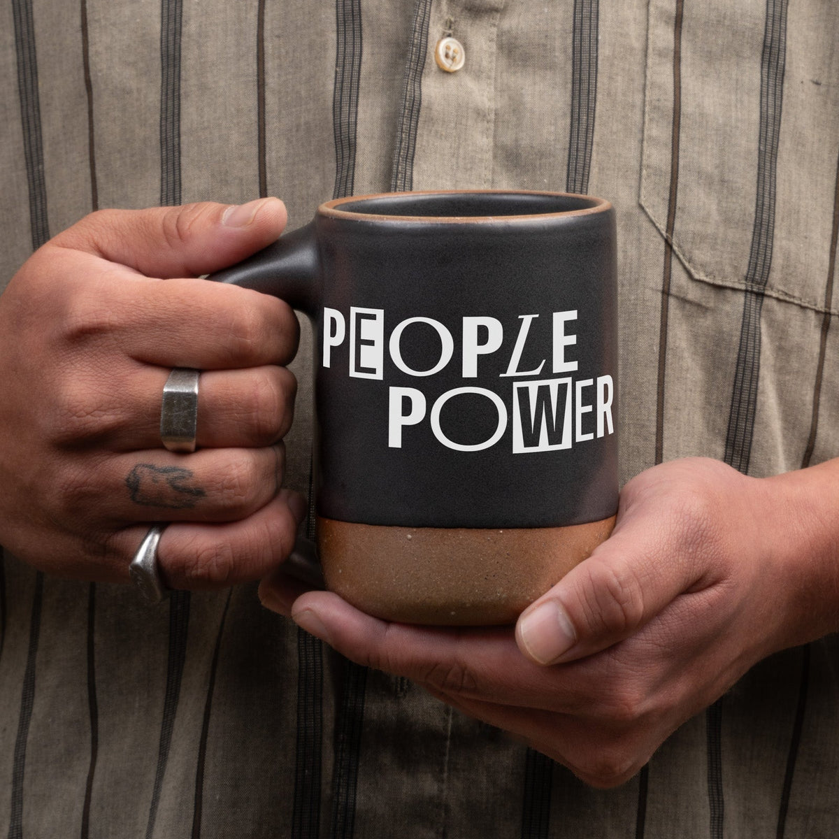 Hands hold up a ceramic mug in a graphite black color with an unglazed base and rim, with a decal in white that reads 'PEOPLE POWER', against a neutral striped button up shirt.