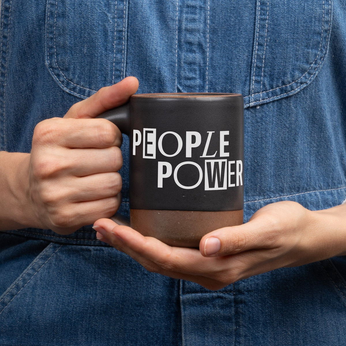 Hands hold up a ceramic mug in a graphite black color with an unglazed base and rim, with a decal in white that reads 'PEOPLE POWER', against a neutral striped button up shirt.