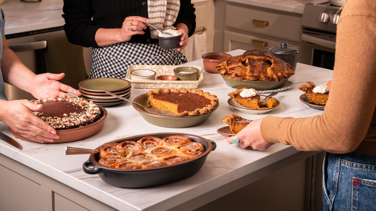 A kitchen scene with multiple baked goods on a countertop, including pies, cakes, and cinnamon rolls. Several people are visible from the shoulders down, serving and holding food items.