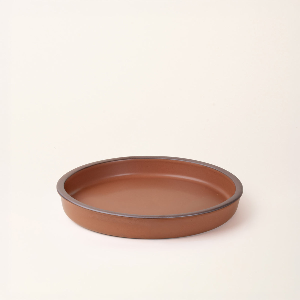 A shallow round ceramic baking dish in a matte terracotta color featuring an unglazed rim, photographed at an angle against a cream background.