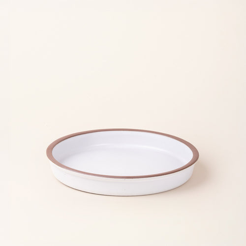 A shallow round ceramic baking dish in a matte cool white color featuring an unglazed rim, photographed at an angle against a cream background.