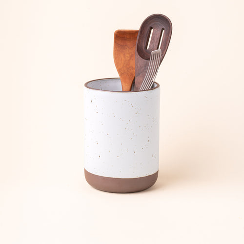 A cylindrical ceramic utensil crock and an unglazed base, in a cool white color, with various kitchen utensils inside