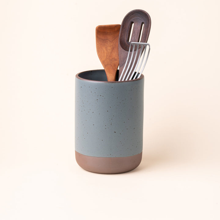 A cylindrical ceramic utensil crock and an unglazed base, in a blue-grey color, with various kitchen utensils inside