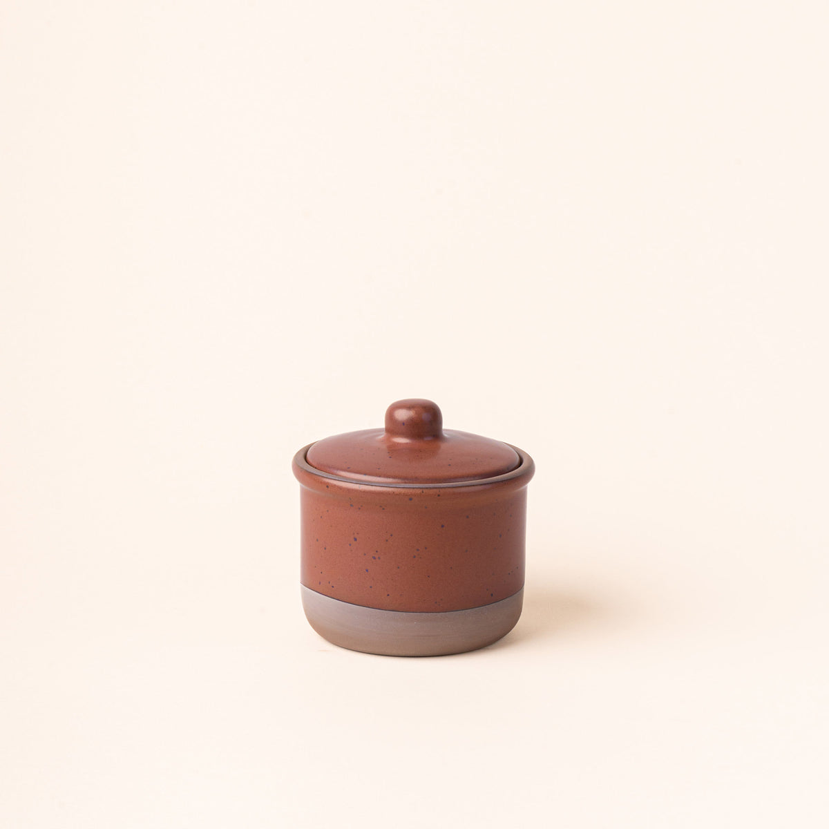 A short ceramic lidded jar in a cool terracotta color with an unglazed base.