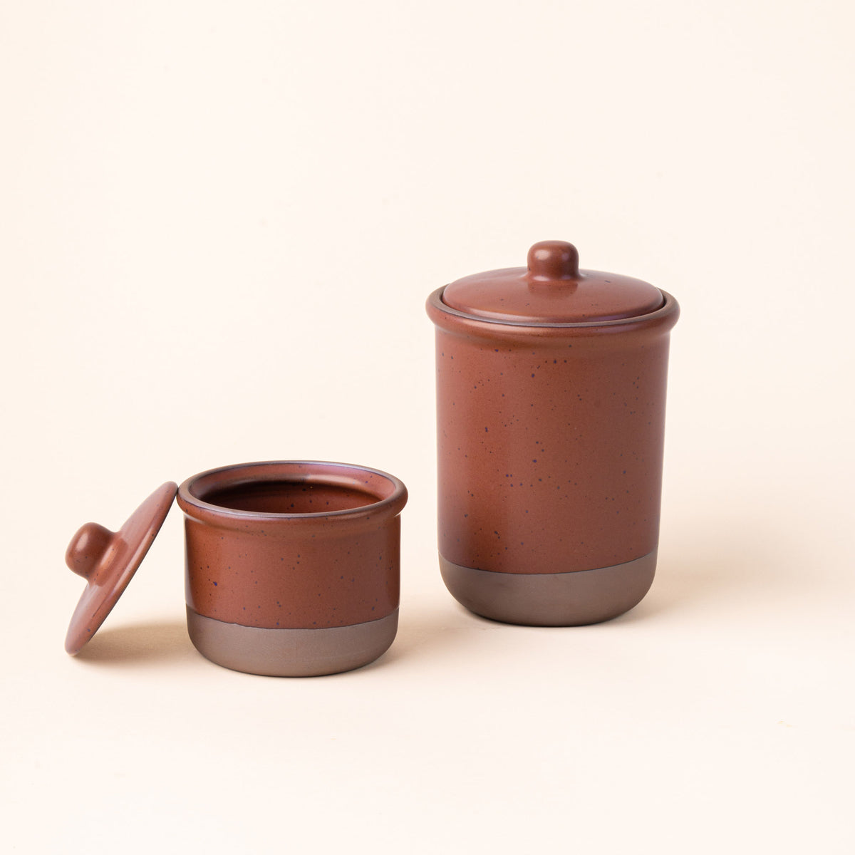 A small and tall ceramic lidded jars in a cool terracotta color with an unglazed base.