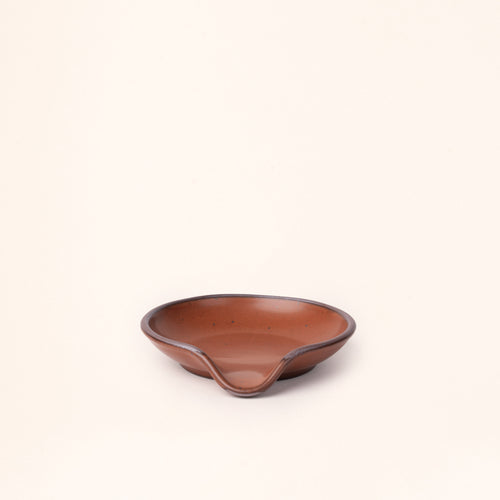 A ceramic spoon rest with a curved notch in a cool terracotta color with an unglazed rim.
