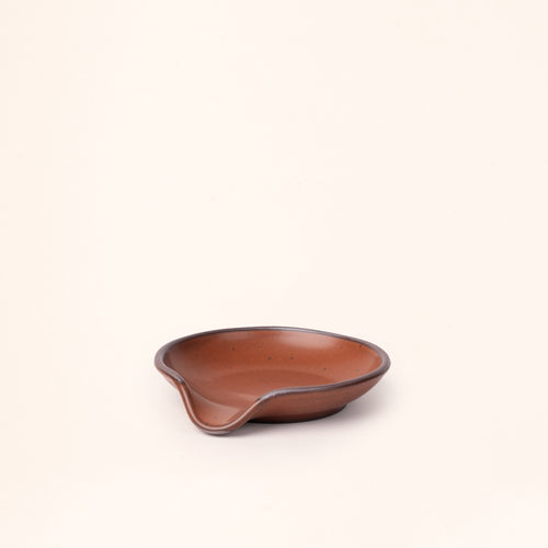 A side view of a ceramic spoon rest with a curved notch in a cool terracotta color with an unglazed rim.