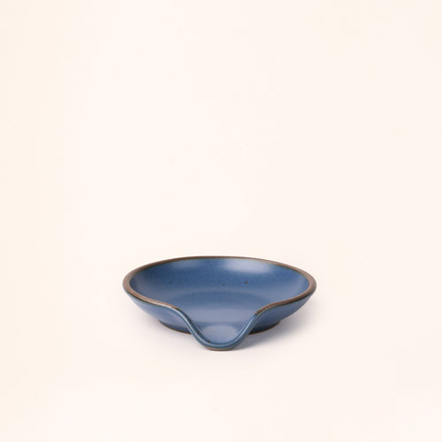 A ceramic spoon rest with a curved notch in a grounded blue color with an unglazed rim.