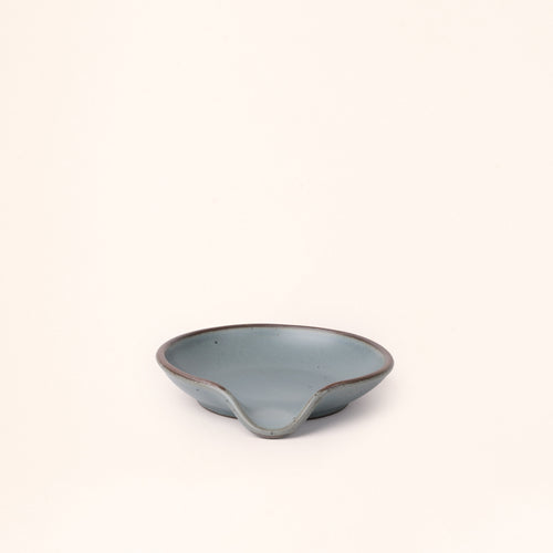 A ceramic spoon rest with a curved notch in a blue-grey color with an unglazed rim.
