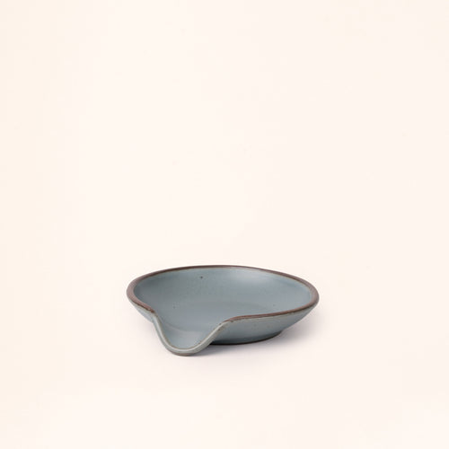 A side view of a ceramic spoon rest with a curved notch in a blue-grey color with an unglazed rim.