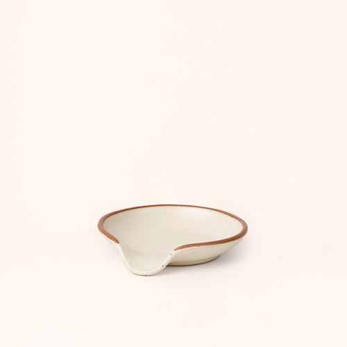 A side view of a ceramic spoon rest with a curved notch in a warm off-white color with an unglazed rim.