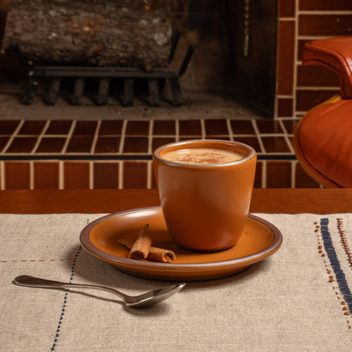 An earthy orange ceramic cup and plate with a creamy drink and cinnamon sticks on a linen tablecloth.