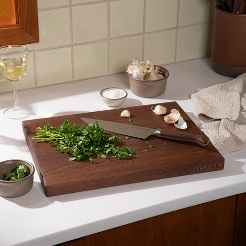 A medium walnut cutting board, with a chef's knife and cilantro being minced with garlic and salt nearby.