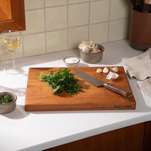 A cherry walnut cutting board, with a chef's knife and cilantro being minced with garlic and salt nearby.