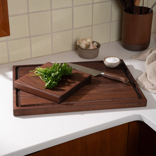 A small square walnut cutting board on top of a larger one with a juice groove, with a chef's knife and cilantro to mince with garlic and salt nearby.