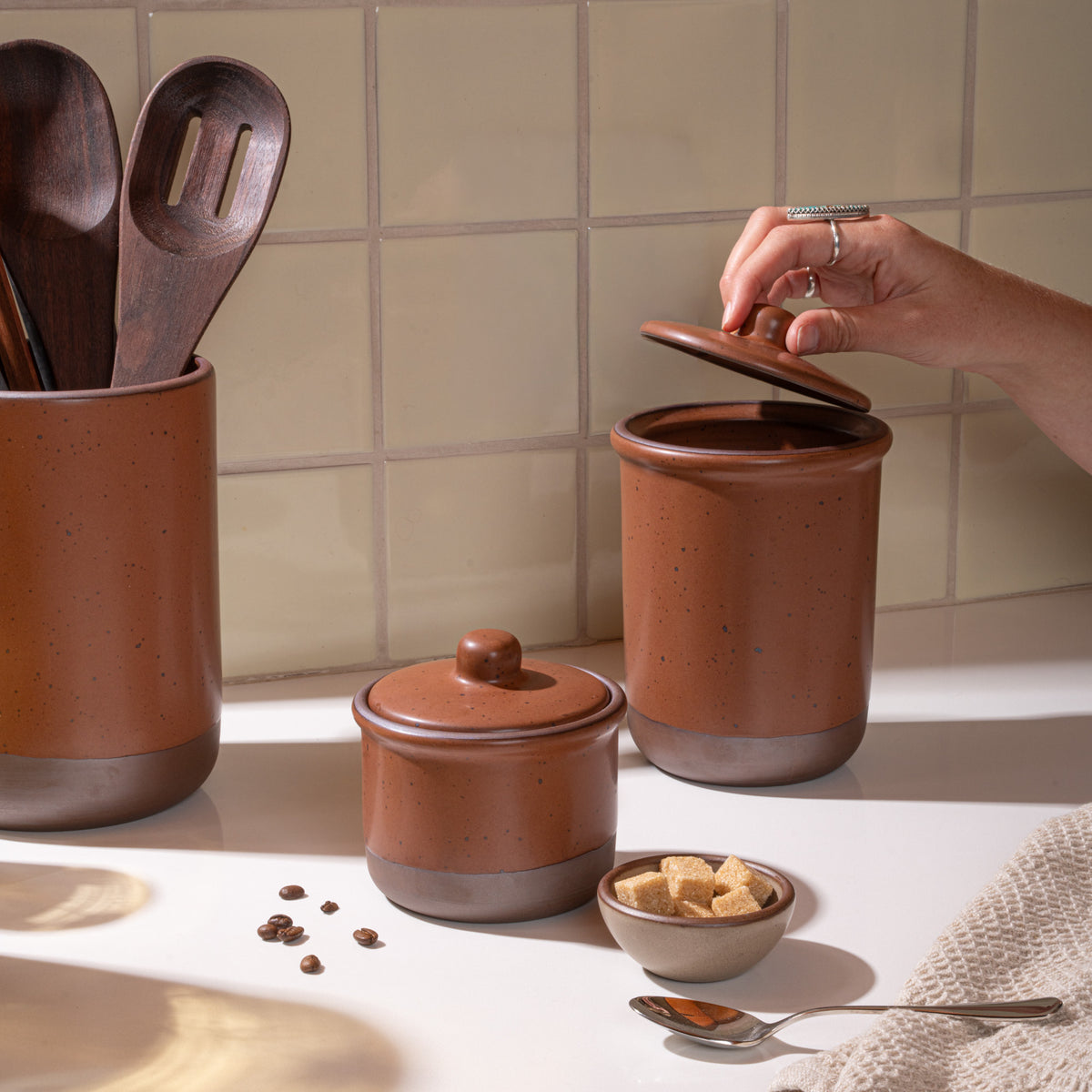 Ceramic utensil crock, small jar and tall jar in a kitchen in a cool terracotta color with kitchen accessories, and a hand opening the jar.
