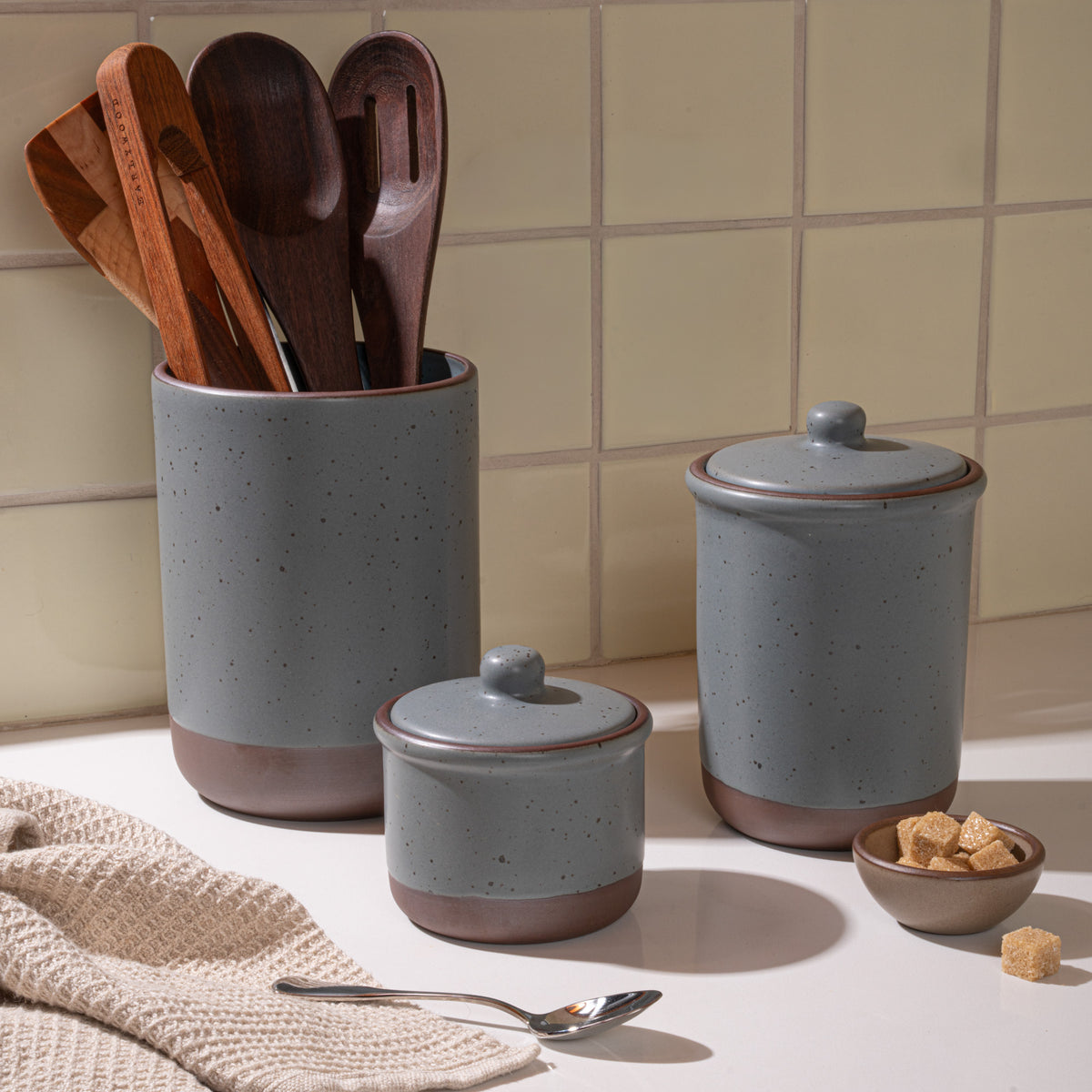 Ceramic utensil crock, small jar and tall jar in a kitchen in a blue-grey color with kitchen accessories.