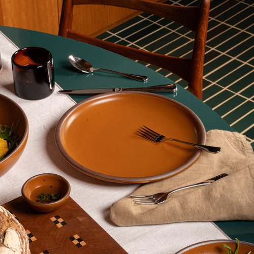 A ceramic dinner plate in an earthy orange glaze, styled on a teal table with silver cutlery, a beige napkin, and matching small bowls in a dining setting.