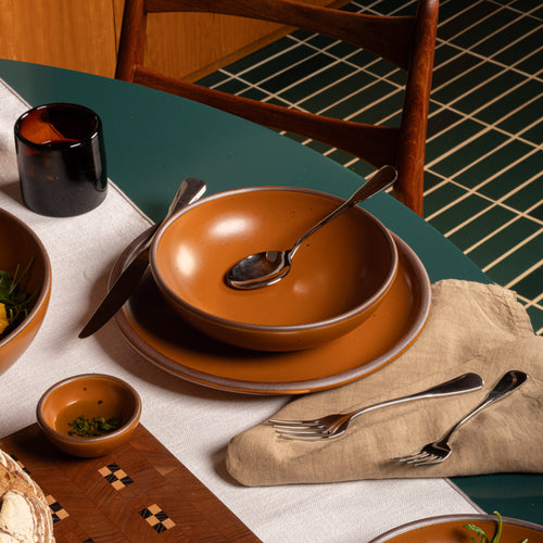 Earthy orange ceramic shallow bowl and dinner plate with silver utensils on a green dining table, surrounded by orange serving dishes.