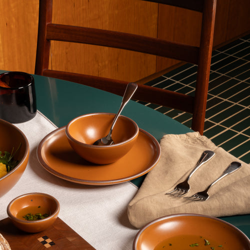 A coordinated earthy orange  dinnerware set arranged on a dining table, including a large plate, matching bowl and saucer, smaller bowls, silver forks on a beige napkin, and a dark mug. A wooden chair and tiled floor are visible in the background.