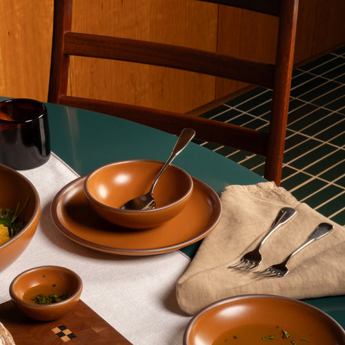 earthy orange ceramic dinnerware set on a dining table, including a bowl, plate, spoon, forks on a napkin, and other bowls, styled with a white runner, wooden chair, and green tiled floor.