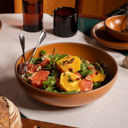 Earthy orange ceramic shallow serving bowl filled with citrus salad, greens, mint, and nuts on a green table.