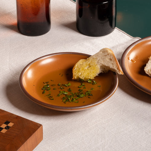 Earthy orange ceramic plate with a bowl edge with olive oil, herbs, and a piece of bread on a linen tablecloth.