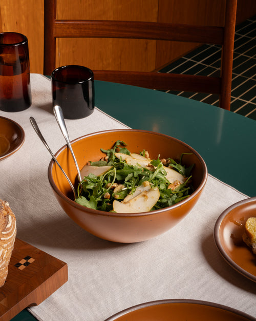 An earthy orange ceramic bowl filled with a green salad and sliced pear, set on a dining table with matching plates, dark amber glasses, and a wooden chair in the background.