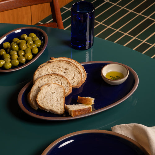 Medium oval ceramic platter in a glossy dark blue color with sliced bread and a small bowl of olive oil on a dark green table.