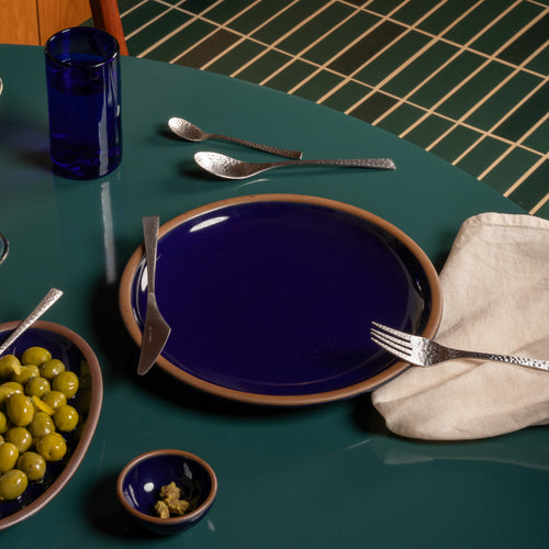 A dark glossy blue ceramic dinner plate with a brown rim, a matching small bowl, a blue glass, and silver cutlery on a dark green tiled table.