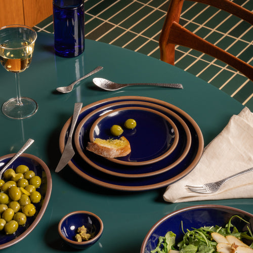 Glossy dark blue ceramic plates with bread and olives, silver utensils, and glassware arranged on a green dining table.