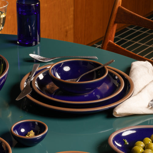 Deep glossy blue ceramic dinnerware with exposed clay rims arranged on a teal table with silverware, a white napkin, and small serving dishes.