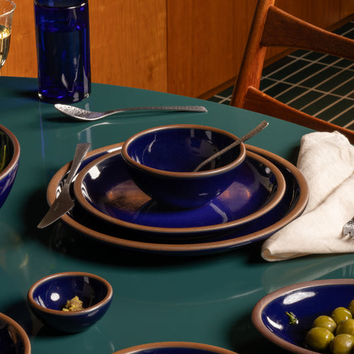 A set of deep glossy blue ceramic dinnerware of a bowl and two plates, with a contrasting brown rim, arranged on a teal table alongside silver cutlery, a white napkin, and green olives.