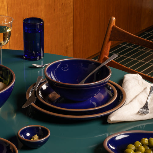 A set of deep glossy blue ceramic tableware, including bowls, plates, and a drinking glass, styled on a teal table with silver cutlery and a white napkin.