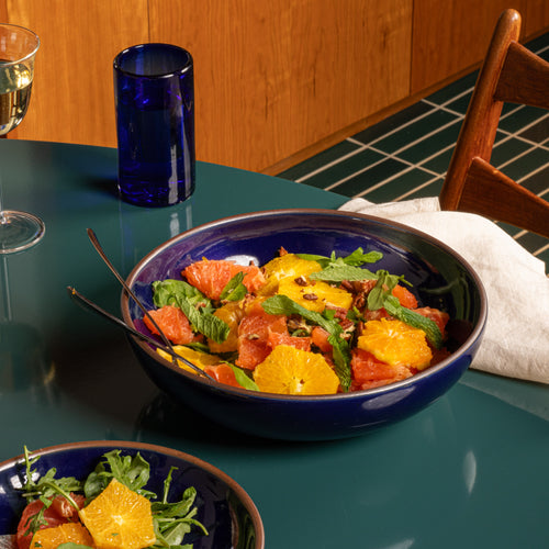 Glossy dark blue ceramic shallow serving bowl filled with citrus salad, greens, mint, and nuts on a green table.
