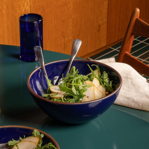 A dark glossy blue ceramic bowl filled with a salad of arugula and pear slices, set on a teal table next to a blue glass and a wooden chair.